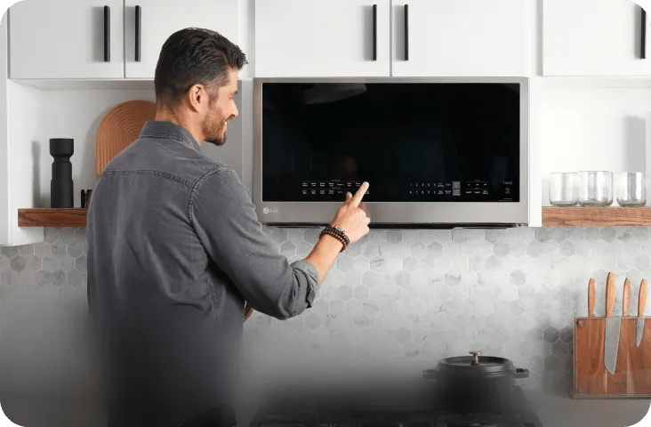 Person standing at a kitchen counter touches the glass control panel of an LG over-the-range microwave mounted below white cabinets and shelves.