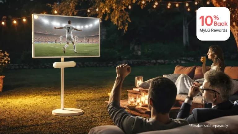 A group of friends gather around a StanbyME 2 Max and speaker in a cozy backyard seating area, watching a soccer match. “10% Back MyLG Rewards” badge.