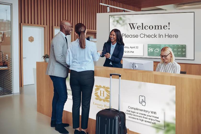 A hotel receptionist greets guests. LG signage behind and on the check-in desk displays hotel information.