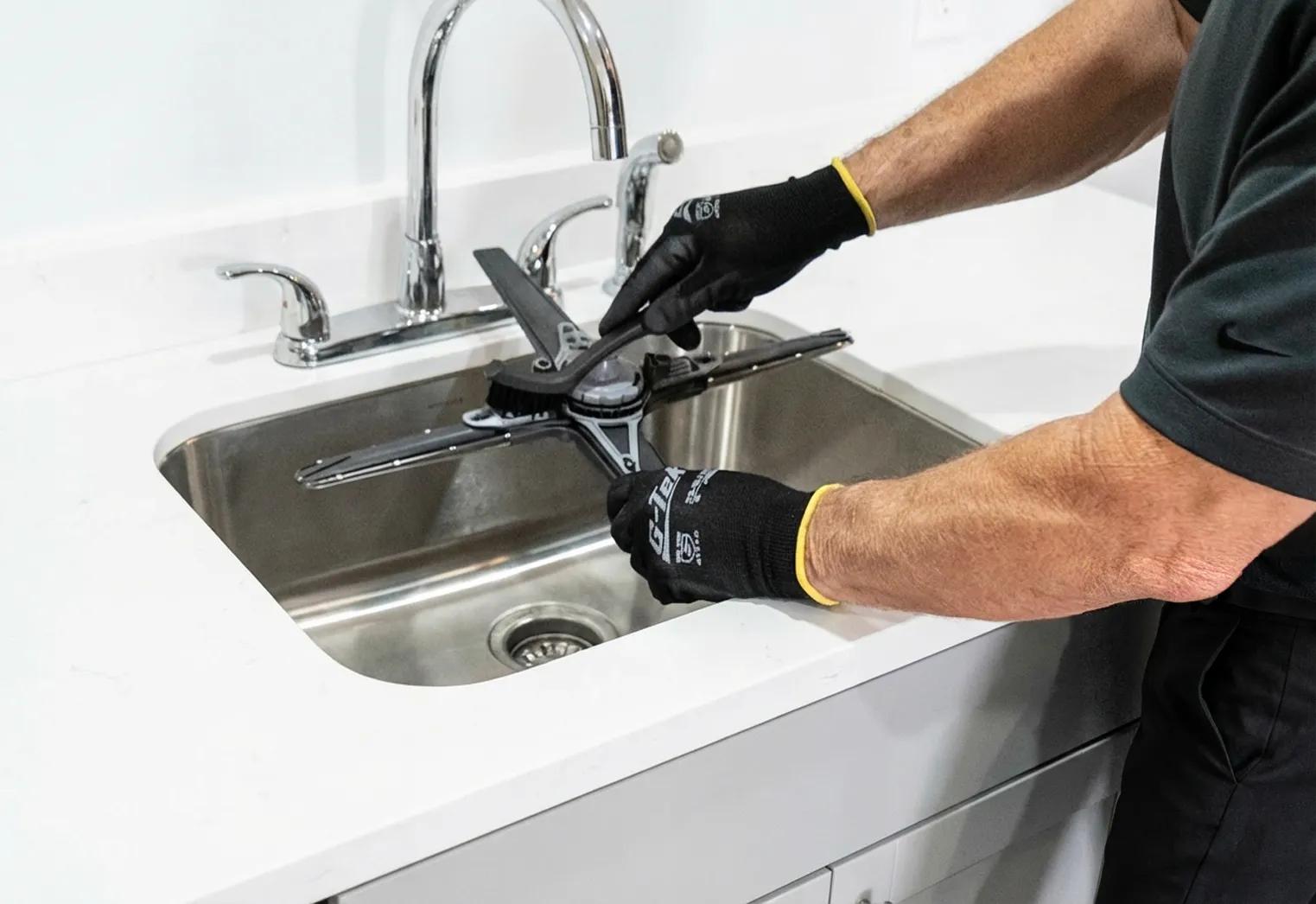 An LG technician wearing black protective gloves cleaning a removable spray arm component from an LG dishwasher in a stainless steel kitchen sink.