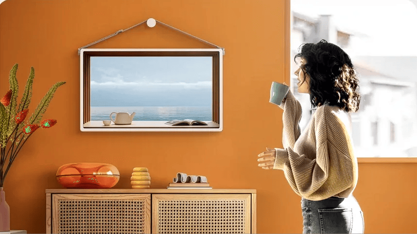 LG StanbyME 2 displayed as a digital picture frame on an orange wall, showing a coastal landscape, with a woman standing nearby holding a cup.