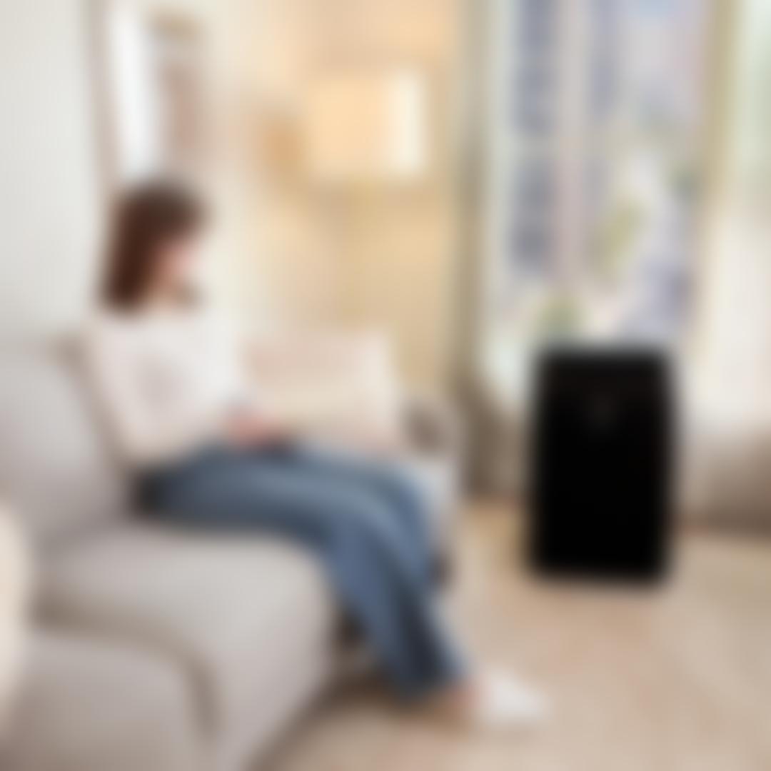 Woman reading on a sofa in a bright city apartment with an LG black portable air conditioner vented through a window overlooking an urban street.