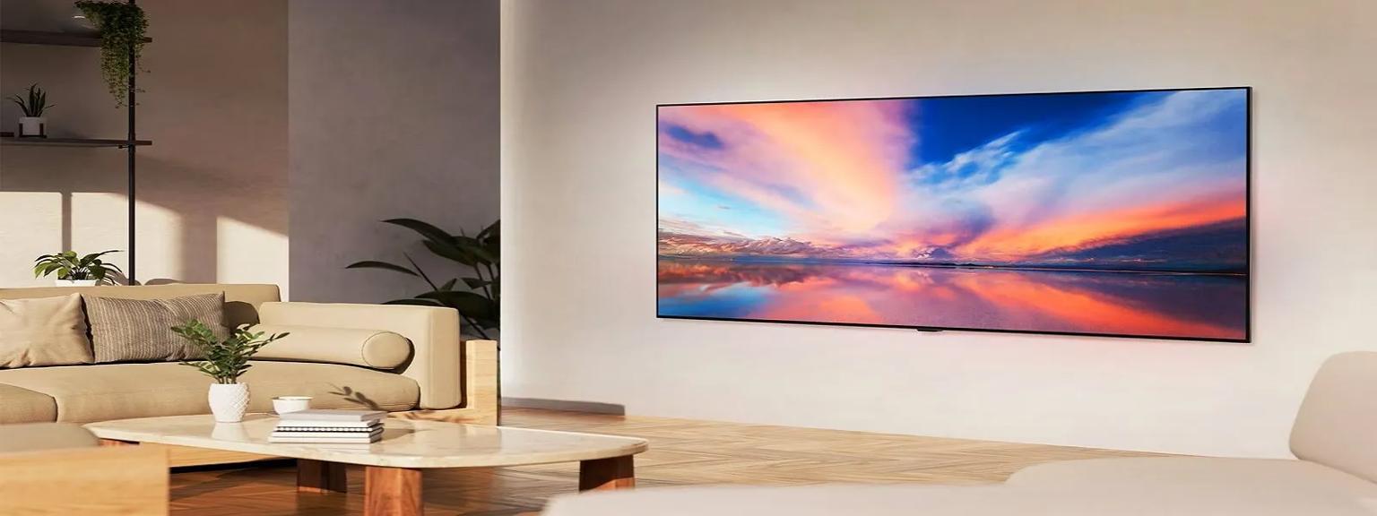 A modern living room with a large wall-mounted TV showing a vivid sunset over a calm lake, cozy beige furniture, a marble coffee table with a plant, and soft natural light filling the space.