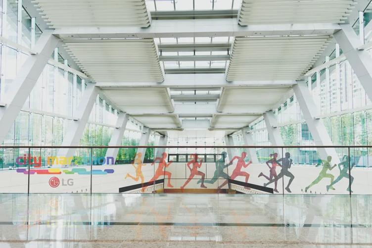 An upper story balcony with a large central open-air space in a classy modern building. The railing for the balcony has Transparent DVLED Film displaying colorful silhouettes of runners, with the words 'City Marathon' and the LG logo to their left.