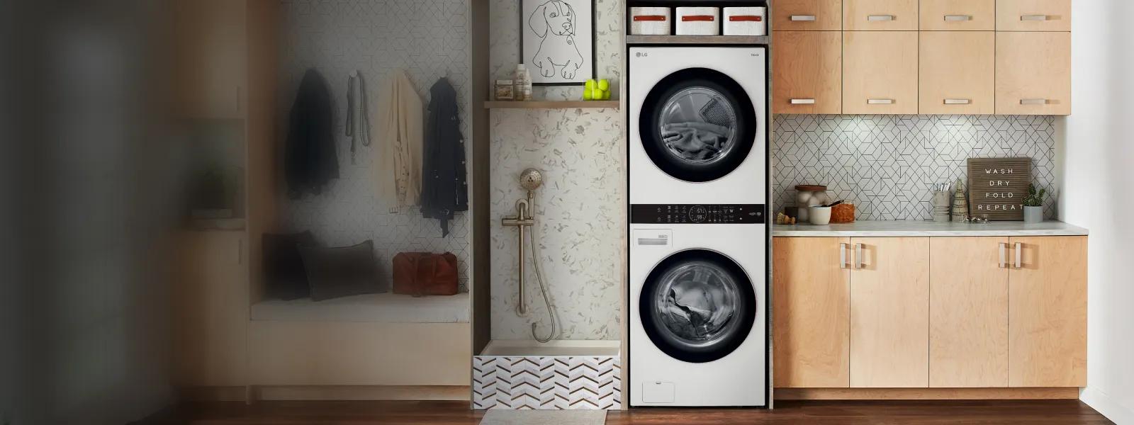 Modern laundry room interior featuring stacked white LG washer-dryer with black drums, marble tile wall with hanging garments and accessories on left, light wood cabinetry with storage and 'Wash Dry Fold Repeat' sign on right.