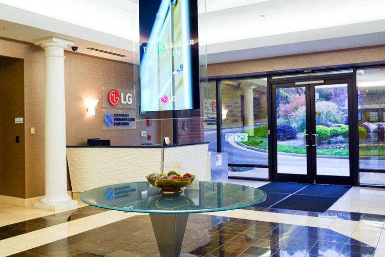 The foyer of the LG Atlanta Business Innovation Center. The LG Logo is on the wall above a receptionist's desk, and an LG transparent signage thanking guests for visiting is hanging over a small end table in the middle of the space. The space is classy and brightly lit, with large windows on either side of the glass entry doors. 