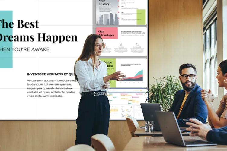 Professionals in an office meeting space. A woman is standing in front of an LG video wall displaying a presentation, talking to three coworkers sitting at the conference table before her. 