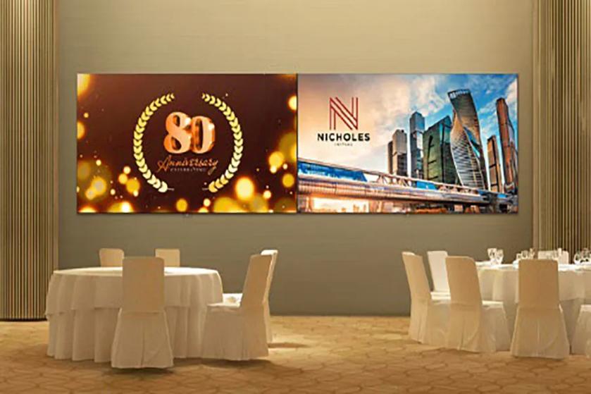 Banquet hall with decorated round tables and two display panels on the wall, one with "80 Anniversary" and the other with "NICHOLES" and a cityscape.