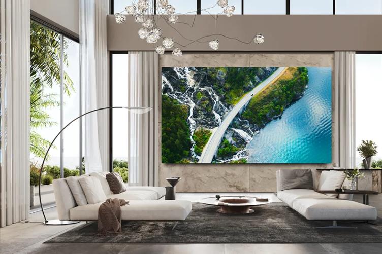 An upscale modern style living room with large windows and pale, chic furniture. An LG residential DVLED display is on one wall, displaying an image of a road curling through a lush coastal area.
