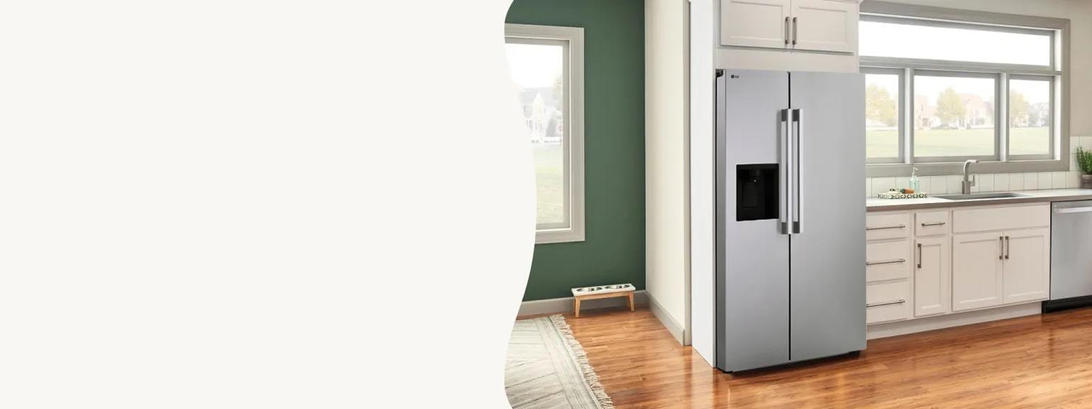 A stainless steel LG side-by-side refrigerator stands next to white cabinetry and a light countertop in a sunlit kitchen. The room features warm wood floors, a green accent wall with a pet bowl station, and a large window overlooking a neighborhood.