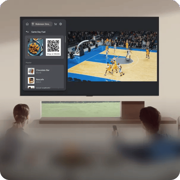 LG TV displaying a basketball game alongside a webOS quick order card showing Game Day Fuel snack items including Chocolate Bar and Nescafe with a QR code to shop on mobile, viewed by two people in a living room