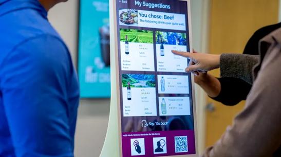 A close-up of an LG touch kiosk in a Business Innovation Center. Several people are standing nearby, cut off partially by the photo frame. Two people are reaching out to touch the display and select options. 