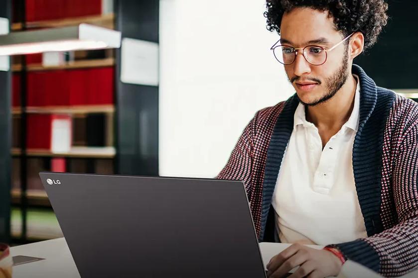 A man with brown curly hair and glasses sits at a table on an LG Ultra PC laptop, showcasing the logo on the front of the laptop. the LG Glance by MiraMetrix l.ogo is in the bottom right corner.
