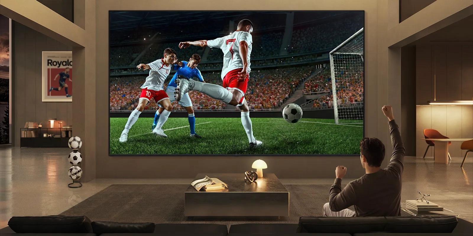LG Micro RGB evo TV with a 100-inch display provides immersive sports viewing, showing a large-scale soccer match with lifelike players and stadium detail filling the screen while a viewer raises one arm in celebration.