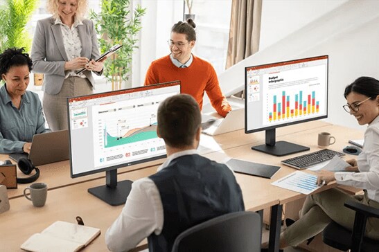 A brightly lit workplace with professionals working at a large open desk. Two workers are using LG monitors, with their displays facing the viewer to show off the boldly colored presentation graphs and charts on their screens. On the opposite side of the table, two other workers use LG laptops to collaborate. A fifth employee stands between them and is looking at one of the laptops and smiling.