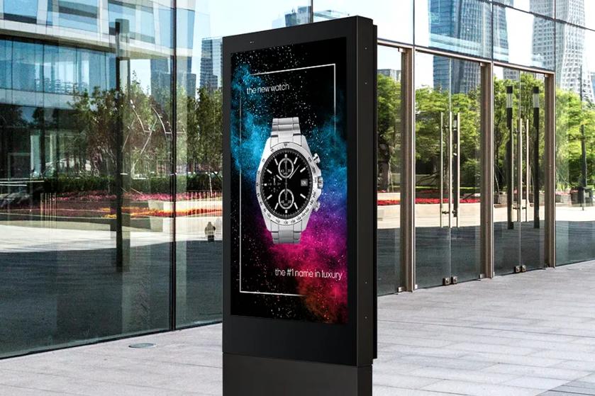 An LG 75XE3C Outdoor Display stands on a sunny city street, outside a reflective glass building. The screen displays a boldly colorful ad for a high quality watch. 