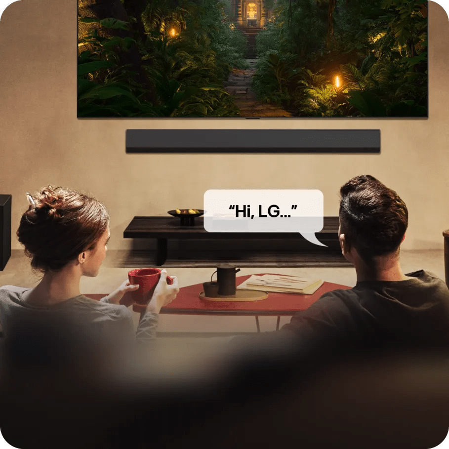 A couple sitting on a couch in a cozy living room watching a large TV showing a lush jungle scene, with a speech bubble next to the man that has the text Hi, LG, indicating voice control.