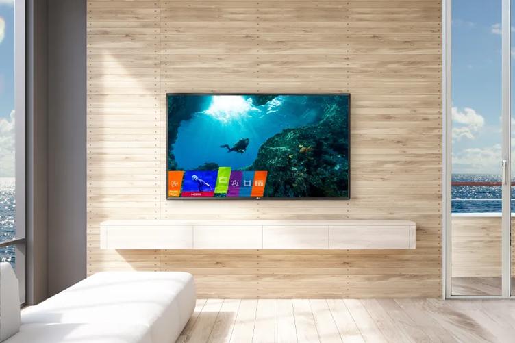 A classy, modern cruise ship cabin. On the wall is an LG Cruise Cabin TV. To its left is a low sofa and a window with an ocean view, and to its right is a glass door leading to a small oceanfront balcony.