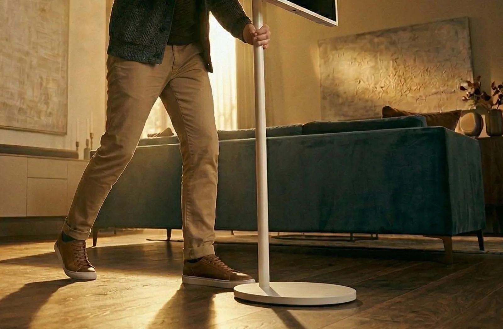 Person rolling LG StanbyME 2 on its wheeled stand across a living room floor.