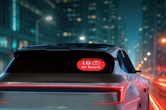 Rear view of a silver car with a red “LG on board” sticker on the window, driving through a brightly lit city at night.