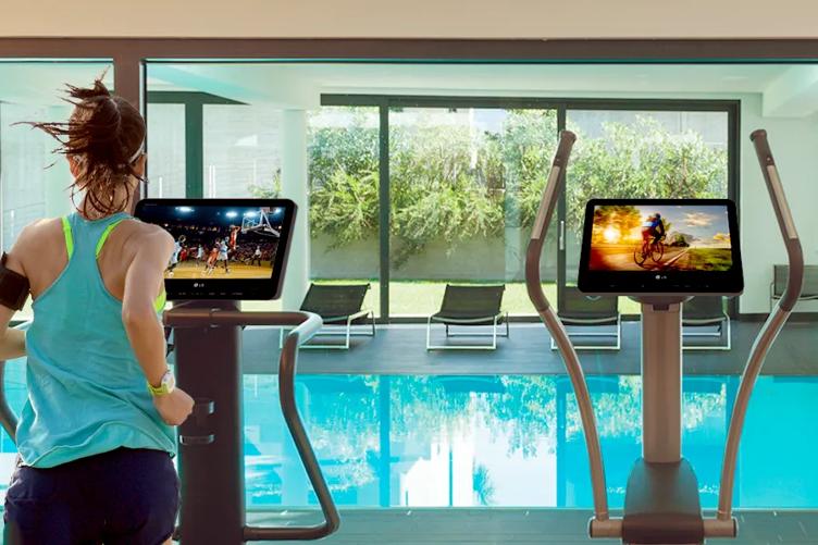 A fitness center with two exercise machines. Beyond them is a large glass window showing a pool room. On the left, a woman runs on a treadmill, using the LG Pro:Centric Smart fitness TV to watch a basketball game. On the right, an elliptical machine sits ready to use, with an LG Pro:Centric Smart fitness TV also mounted to it. The elliptical's TV shows a bicyclist in sports gear riding his bike.