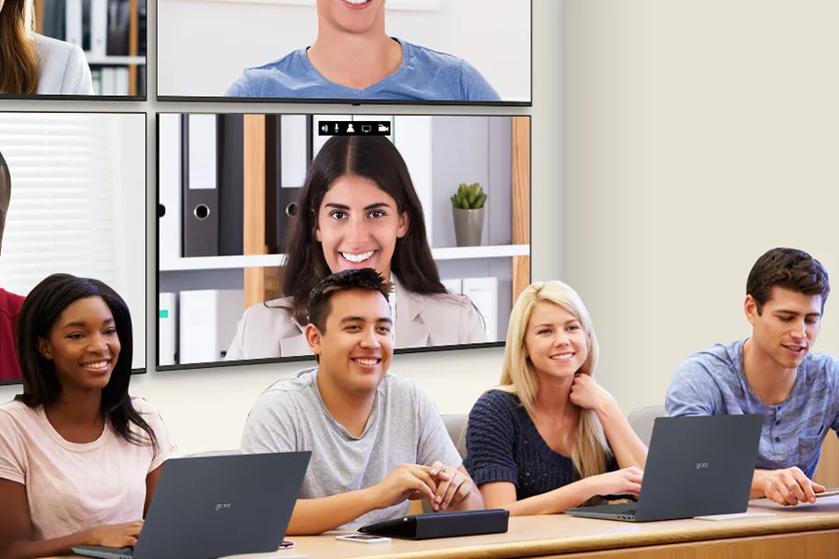 A row of university students sit in a classroom at their desks. Behind them, video wall signage is mounted on the wall, with additional students attending class virtually via video call displayed on the signage.