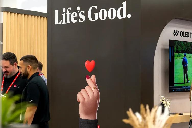 A photograph of the LG HITEC 2025 booth. The main focus of the image is the LG gray wall with Life's Good in white above a print of the LG emoji hand. The hand's fingers are crossed to form a heart, and a cartoon heart floats above them. To the right, an LG TV is just visible inside the booth. To the left, two LG salesmen laughing and smiling are just visible. 