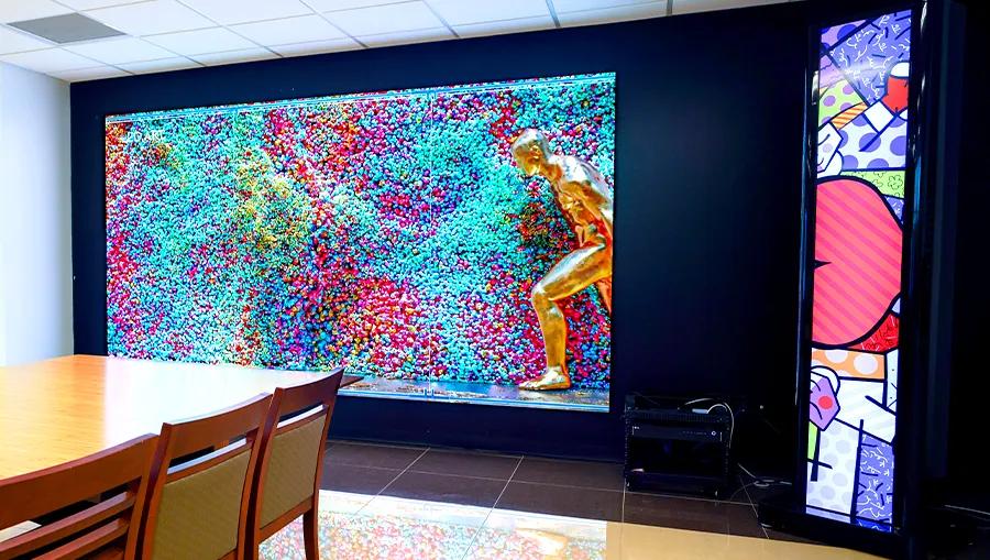 An LG Direct View LED display is mounted on the wall, displaying a bright abstract art. To its right, a piece of LG stretch signage is mounted nearby, displaying vivid and colorful pop art. The light and color from both is reflecting off the floor and the nearby conference table.