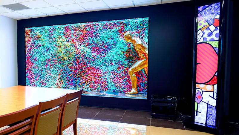 An LG Direct View LED display is mounted on the wall, displaying a bright abstract art. To its right, a piece of LG stretch signage is mounted nearby, displaying vivid and colorful pop art. The light and color from both is reflecting off the floor and the nearby conference table.