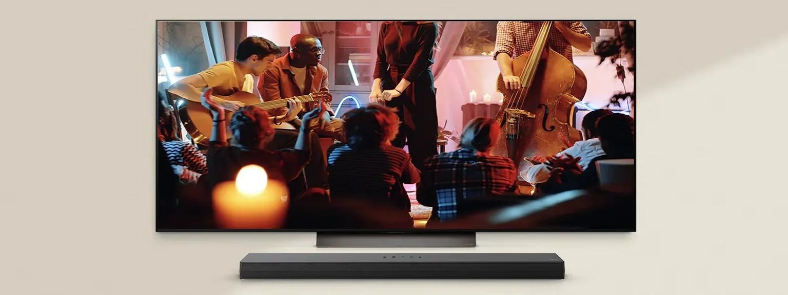An LG TV showcases a cozy live music performance, featuring musicians playing guitar and double bass in a warmly lit setting. Below the LG TV, an LG Soundbar is positioned, enhancing the immersive atmosphere.