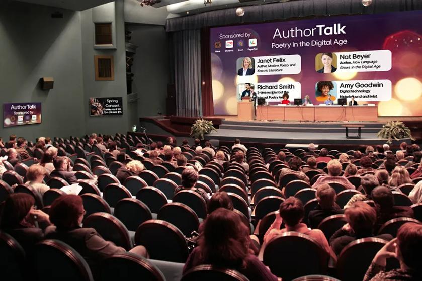 A large university theater space, with an author talk happening on stage. A DVLED display forms the backdrop of the stage, showcasing the event title and author information, and signage along the walls display additional event information. 