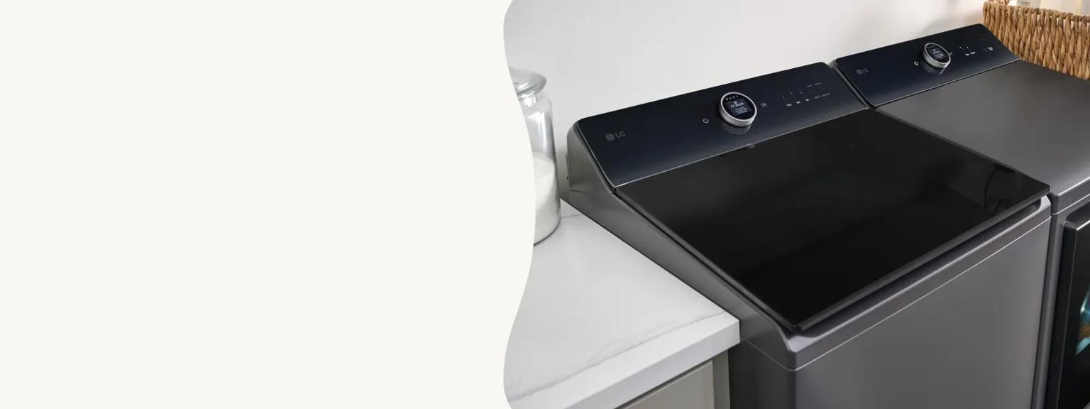 A high-angle, close-up shot of a modern LG top-load washer and matching dryer in a sleek matte charcoal finish. The appliances feature dark glass lids and minimalist digital control dials on the back consoles, positioned next to a white marble countertop and a wicker basket.