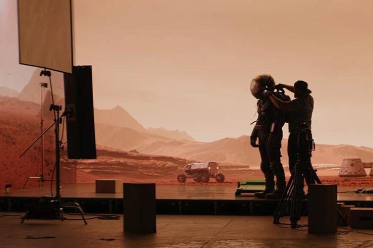 A studio hand adjusts an actor's astronaut helmet. Behind them is a large LG DVLED video wall, showing a red dusty planet landscape for the movie's backdrop.