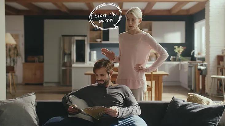 In a modern smart kitchen, a woman uses a voice command to start the washer with LG ThinQ AI, while a man reads on the sofa in the background.