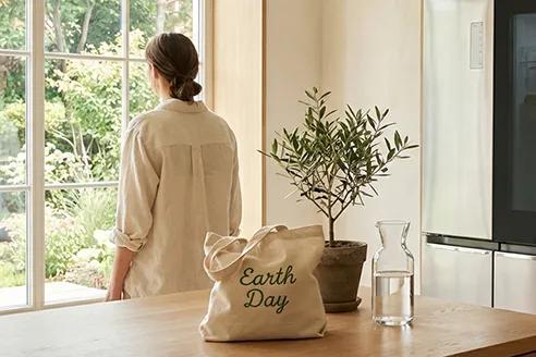 Woman standing near a bright window beside an 'Earth Day' tote bag, a small potted olive tree, and a glass carafe on a wooden countertop.