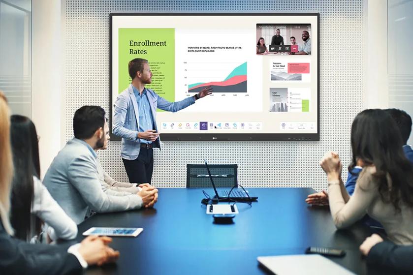A faculty member gestures at a LG CreateBoard on a meeting room wall as he gives a presentation on enrollment rates. Other staff are seated at a conference table, watching his presentation with interest.