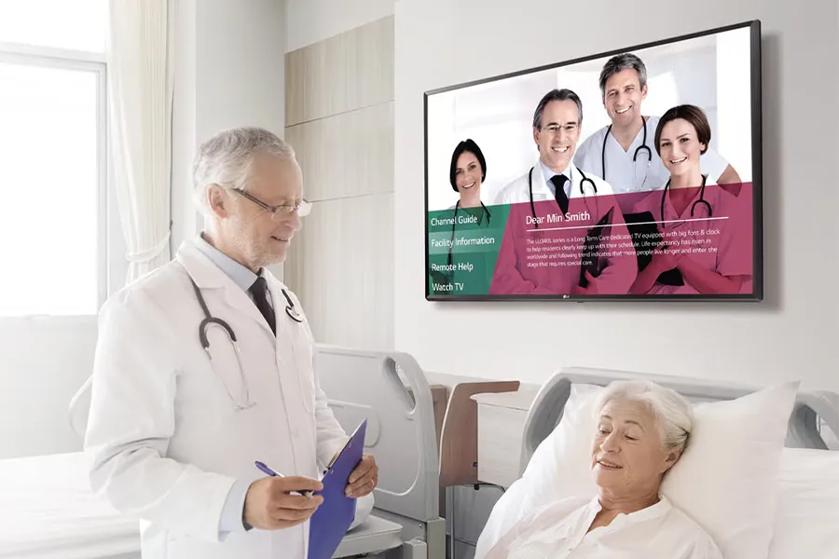 A doctor in a hospital room stands beside a bed with an elderly patient, with a TV screen displaying healthcare professionals and text.