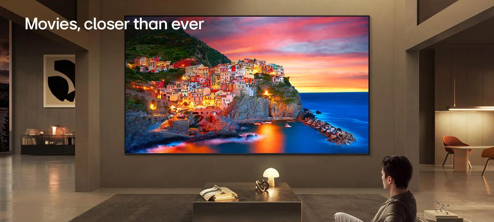 LG Ultra Big TV is wall-mounted in a spacious living room, where an expansive display shows a vibrant coastal village at sunset, illustrating how a larger screen delivers an immersive and cinematic viewing experience.