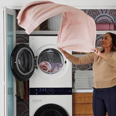 A woman in a tan sweater dynamically shaking out a large pink bath towel in front of a white LG WashTower stacked laundry unit.