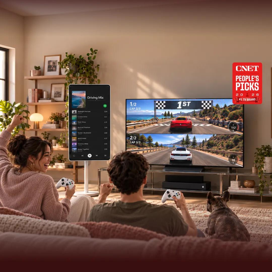A couple plays a split-screen racing game on a large LG TV, controllers in hand, celebrating a 1st-place finish, with a French bulldog watching beside them on the couch. A portable smart display shows a "Driving Mix" playlist nearby. Cozy living room setting with plants and shelving. A "CNET People's Picks 2026 #1 TV Brand" badge appears in the upper right.