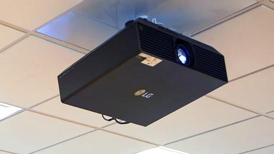 An LG ProBeam projector mounted on the ceiling of the LG Atlanta Business Innovation Center. The lens is lit up, indicating that it's projecting images. 