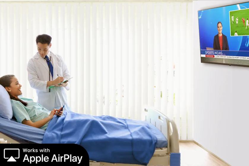 A woman is sitting in a hospital bed with a doctor standing beside her. The doctor is looking at a clipboard. The patient is smiling as she uses her smartphone to cast a news show to the LG Pro:Centric SMART hospital TV on the wall before her.