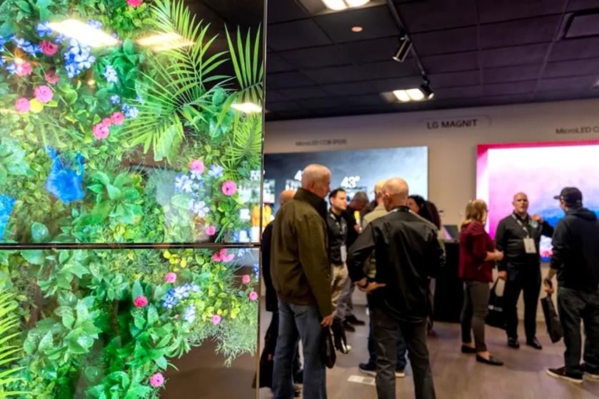 To the left side of the image, a transparent OLED signage display shows bright green foliage dotted with vividly colored flowers. In the background behind it, to the right, guests mingle and admire LG digital displays and other solutions in the Business Innovation Center.