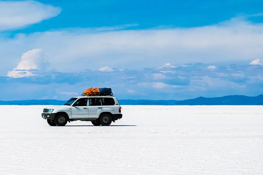 A white SUV with luggage on the roof drives on a vast salt flat under a clear blue sky.