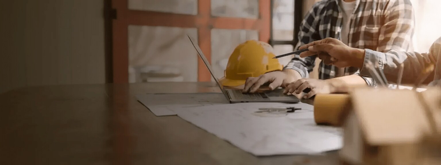 A close up on two builders standing side by side at a workbench with blueprints spread out in front of them. One points at a laptop screen with a pencil.