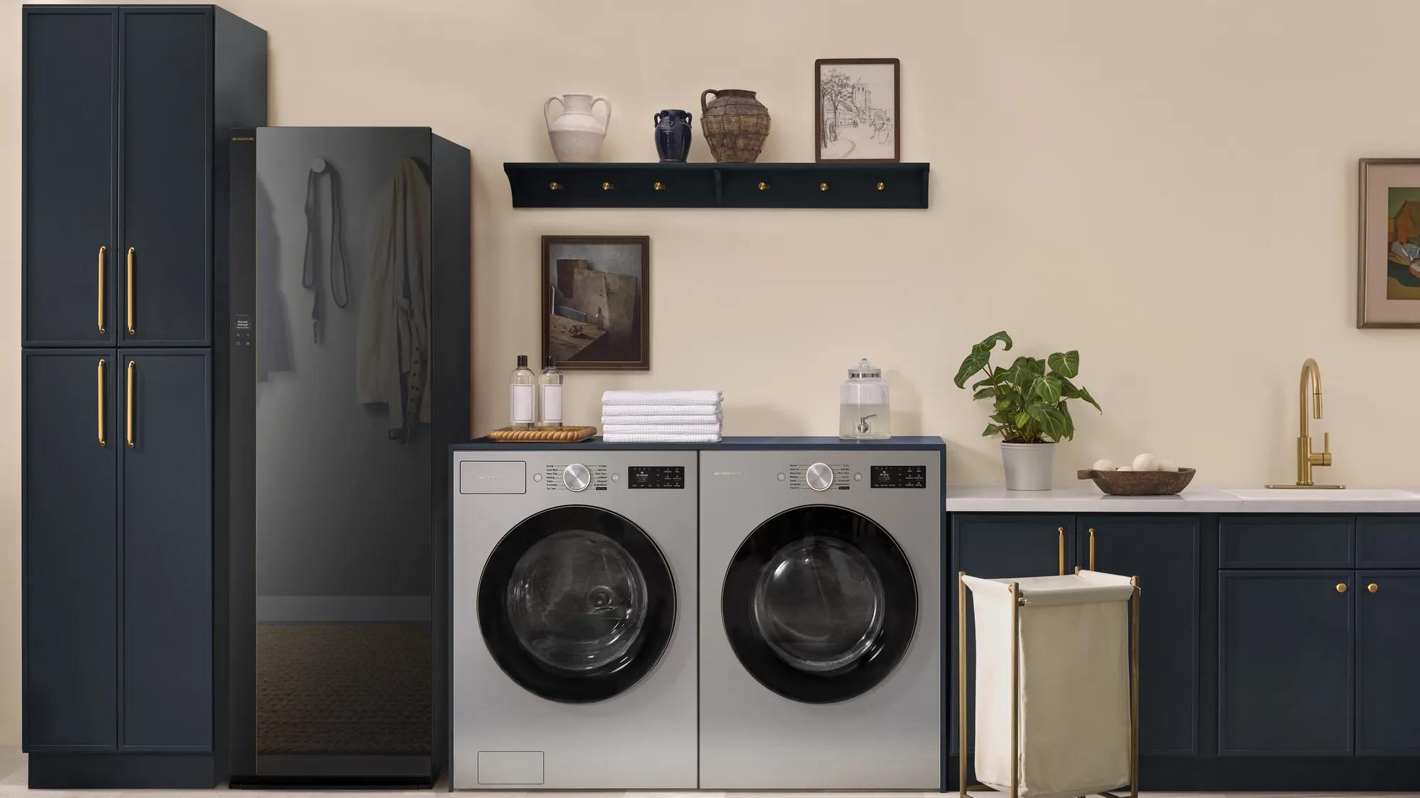 A styled laundry room with cream walls and navy cabinets with brass hardware. On the left, a tall clothing care cabinet with a dark tinted glass door shows a coat and a bag hanging inside. Next to it, a silver front-load washer and dryer sit side by side under a navy countertop with folded white towels, detergent bottles and a glass dispenser. A floating shelf with ceramic vases and framed art hangs above, and a brass faucet, a potted plant and a canvas laundry hamper sit on the right.