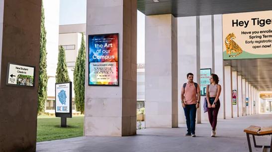 Two students walk across campus, surrounded by various LG signage advertising university events. 
