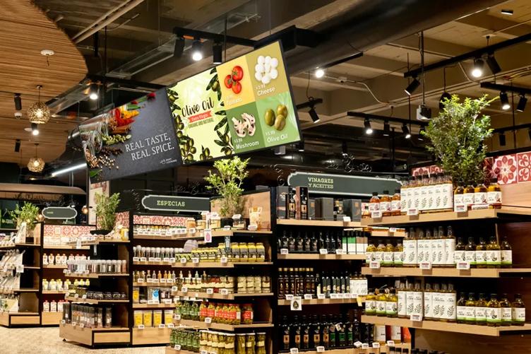 upermarket aisle with shelves of oils, jars, and condiments; overhead digital advertisements for food items.