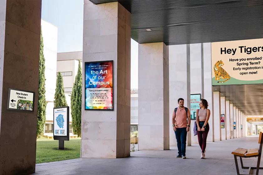 Two students walk across campus, surrounded by various LG signage advertising university events. 
