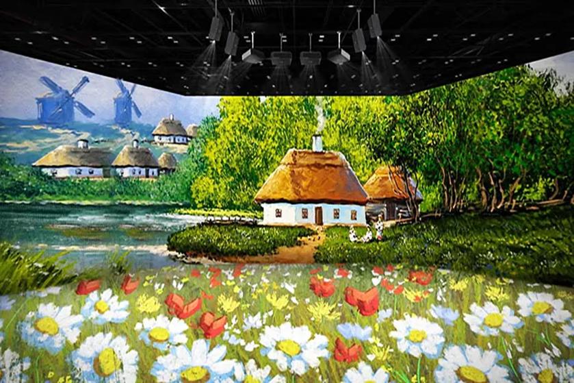 The image showcases a vibrant, digitally projected artwork within an indoor space, suggesting a 3D immersive environment. In the foreground, there is a meadow filled with daisies and scattered red poppies. A thatched-roof cottage with white walls and brown doors sits to the right, surrounded by lush green trees. Smoke rises from the chimney, suggesting a cozy, inhabited dwelling. In the background, multiple thatched-roof cottages are nestled in a landscape with a serene lake and fields. Two prominent windmills are visible against a blue sky, adding to the pastoral atmosphere. Overhead, several dark ceiling lights suggest an exhibition or digital gallery setting